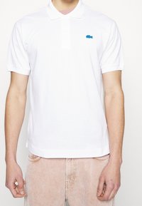 Polo blanc en coton avec un col à trois boutons et des manches courtes, orné d'un logo Lacoste bleu sur la poitrine.