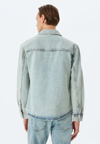 Vaaleansininen farkkutakki, jossa on rento leikkaus, näkyvät saumat ja nappikiinnitys hihansuissa. Mukana klassinen kaulus ja hieman kulunut kangas.