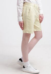 Shorts jaune clair en coton, avec une coupe slim, un devant plat et des poches latérales, assortis à des chaussures blanches et une chemise à motifs.