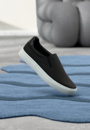 Mustad libisevast materjalist kingad tekstuurse pinnaga, valge kontrastse tallaga, mis on paigutatud sinise vaiba kohale. Minimalistlik disain sujuvate servadega.