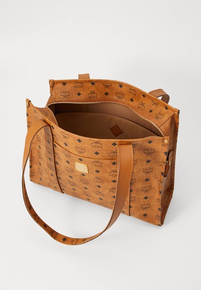 Tan tote bag made from textured material with a patterned design. Features dual handles, a zippered interior, and a decorative logo plate.