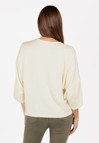 Maglietta oversized di colore crema chiaro con maniche 3/4, scollatura rotonda, texture morbida e vestibilità rilassata, abbinata a pantaloni aderenti di colore verde oliva.