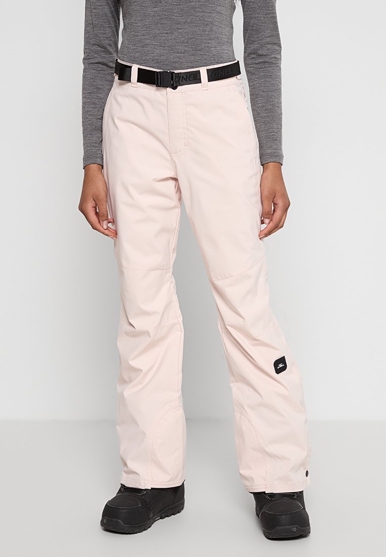 Osoba w jasnoróżowych spodniach narciarskich z czarnym paskiem i czarnych butach śniegowych, stojąca na jednolitym jasnym tle.
