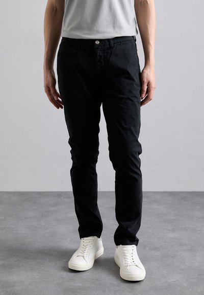 Černé slim fit kalhoty s hladkou texturou a předními kapsami, v kombinaci s bílými teniskami. Oblečení doplňuje jednoduché šedé tričko.