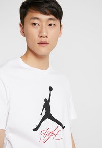 Weißes Baumwoll-T-Shirt mit einer großen schwarzen Silhouette eines Basketballspielers und rotem "Flight"-Text auf der Vorderseite. Rundhalsausschnitt-Design.