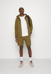 Olive green lightweight jacket with hood, black accents, paired with matching shorts. White t-shirt and beige sneakers complete the look.