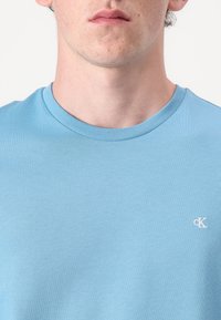 Ανοιχτό μπλε βαμβακερή μπλούζα με στρογγυλή λαιμόκοψη, που διαθέτει ένα μικρό λευκό λογότυπο στην αριστερή πλευρά του στήθους. Λείος υφή.