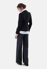 Schwarze Cordjacke mit taillierter Passform, langen Ärmeln und Rückenschlitz, kombiniert mit weit geschnittenen, anthrazitfarbenen Hosen und einem weißen Hemd darunter.