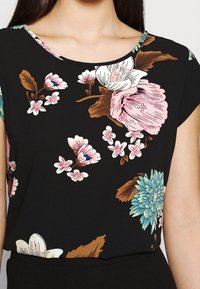 Top noir à fleurs avec de grandes fleurs roses et bleues, des feuilles vertes et un décolleté en forme de U. Fabriqué en tissu léger avec des manches courtes.