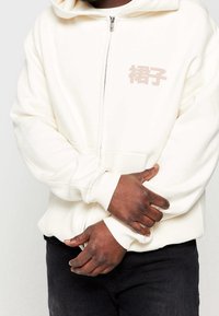 Uomo che indossa una felpa bianca con cerniera e caratteri asiatici sul petto, mani incrociate sopra il basso ventre, e pantaloni neri.