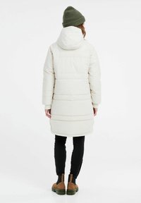 Pružný, středně dlouhý kabát v barvě off-white, s kapucí, žebrovanými manžetami a horizontálním prošíváním. V kombinaci s černými legínami a zelenými botami.