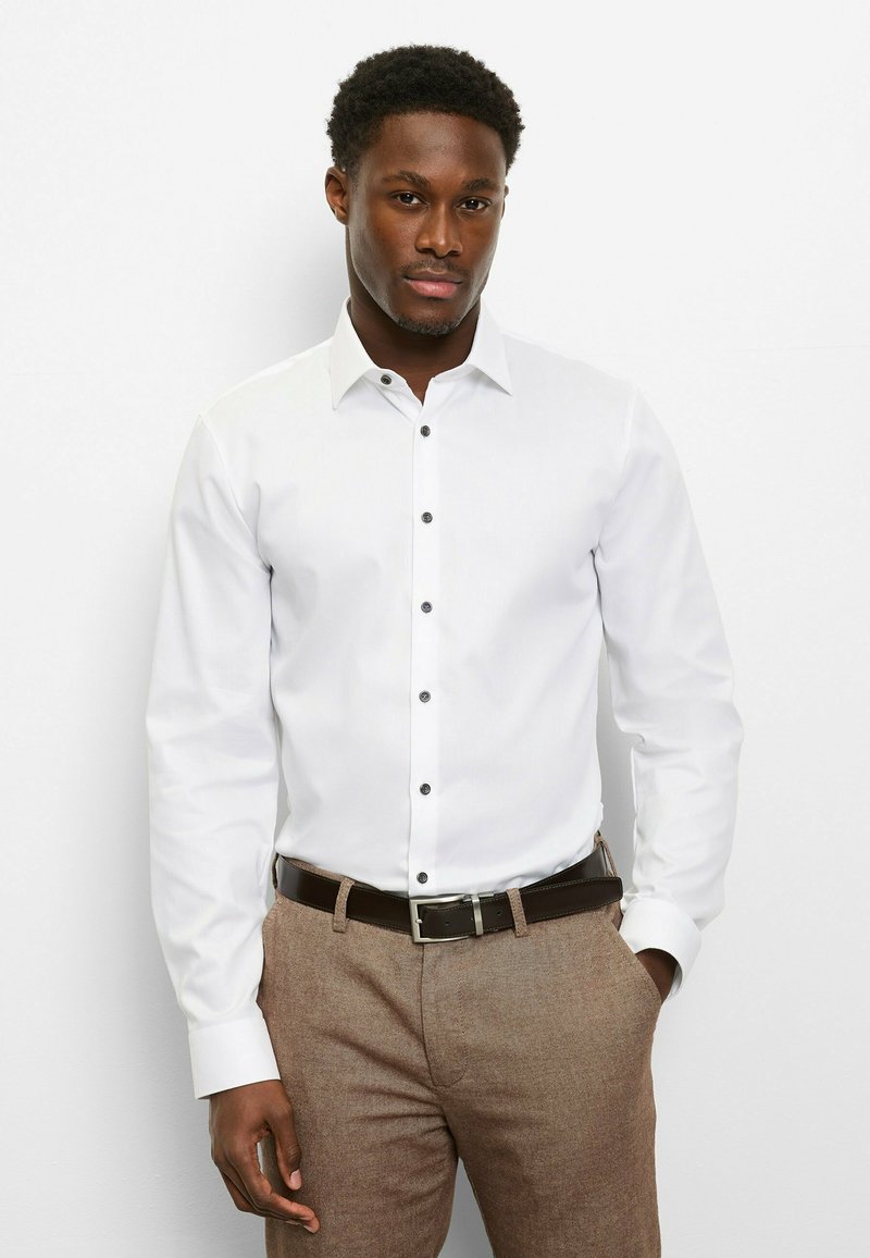 Weißes Langarmhemd mit Knopfkragen und schwarzen Knöpfen, kombiniert mit brauner, strukturierter Hose und braunem Gürtel.