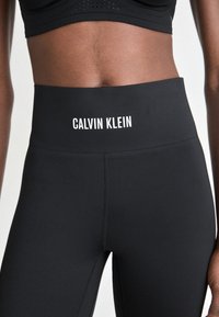Černé legíny s vysokým pasem s bílým logem Calvin Klein na pase, nošené s černým perforovaným sportovním braletem na tmavé pleti.