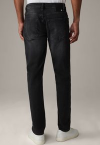 Schwarze Denim-Jeans mit geradem Schnitt, ausgestattet mit Gesäßtaschen, subtiler Textur und einem kleinen Logotag am Bund. Weiße Sneakers sind sichtbar.