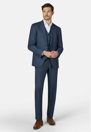 Man in een driedelig marineblauw pak met een wit overhemd en bruine nette schoenen, met handen gevouwen, kijkend naar de zijkant.