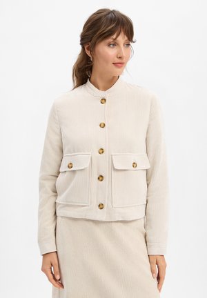 Veste en velours côtelé beige avec un col mandarin, des boutons sur le devant et deux poches poitrine. Possède une texture côtelée et des boutons dorés.