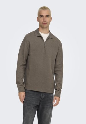 Brun zip-up tröja med krage, strukturerad tyg och långa ärmar, matchad med mörkgrå jeans. Modellen står mot en ljus bakgrund.