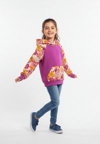 Uśmiechnięta młoda dziewczyna stojąca na jednej nodze, ubrana w niebieskie dżinsy, niebieskie trampki oraz fioletową bluzę z kapturem z różowo-żółtymi kwiatowymi rękawami i kieszenią.