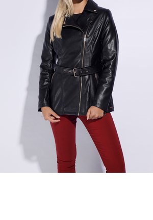 Femme portant une veste en cuir noire ceinturée avec une fermeture éclair diagonale et un pantalon rouge ajusté, debout devant un fond uni clair.