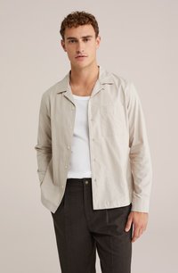 Camicia button-up beige chiaro con colletto aperto, taschino frontale e maniche lunghe, indossata sopra una t-shirt bianca a girocollo e pantaloni scuri.