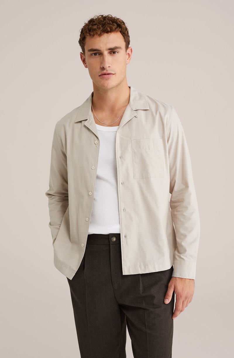 Camicia button-up beige chiaro con colletto aperto, taschino frontale e maniche lunghe, indossata sopra una t-shirt bianca a girocollo e pantaloni scuri.