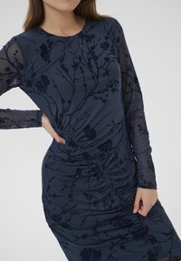Tengeri dressz hosszú áttetsző ujjakkal, virágmintás velúr anyaggal, oldalt fodros részletezéssel és testhezálló sziluettel. Sima textúra, elegáns dizájn.