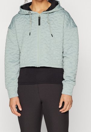 Persoon draagt een lichtblauw, doorgestikt, cropped hoodie met een ritssluiting aan de voorkant en trekkoorden, gecombineerd met zwarte leggings en een zwarte onderlaag.