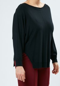 Maglione nero a maglia con scollatura a barca, vestibilità oversize e polsini a costine. L'orlo presenta spacchi laterali. Indossato con pantaloni rossi.