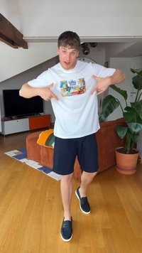 Jeune homme en t-shirt blanc à motif graphique et short noir pointant son t-shirt, debout sur un sol en bois dans un salon moderne.