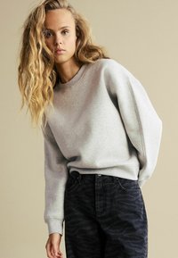 Grauer Baumwoll-Sweatshirt mit langen Ärmeln, Rundhalsausschnitt und Rippbündchen, kombiniert mit dunklen, gemusterten Jeans im Tigerstreifen-Design.