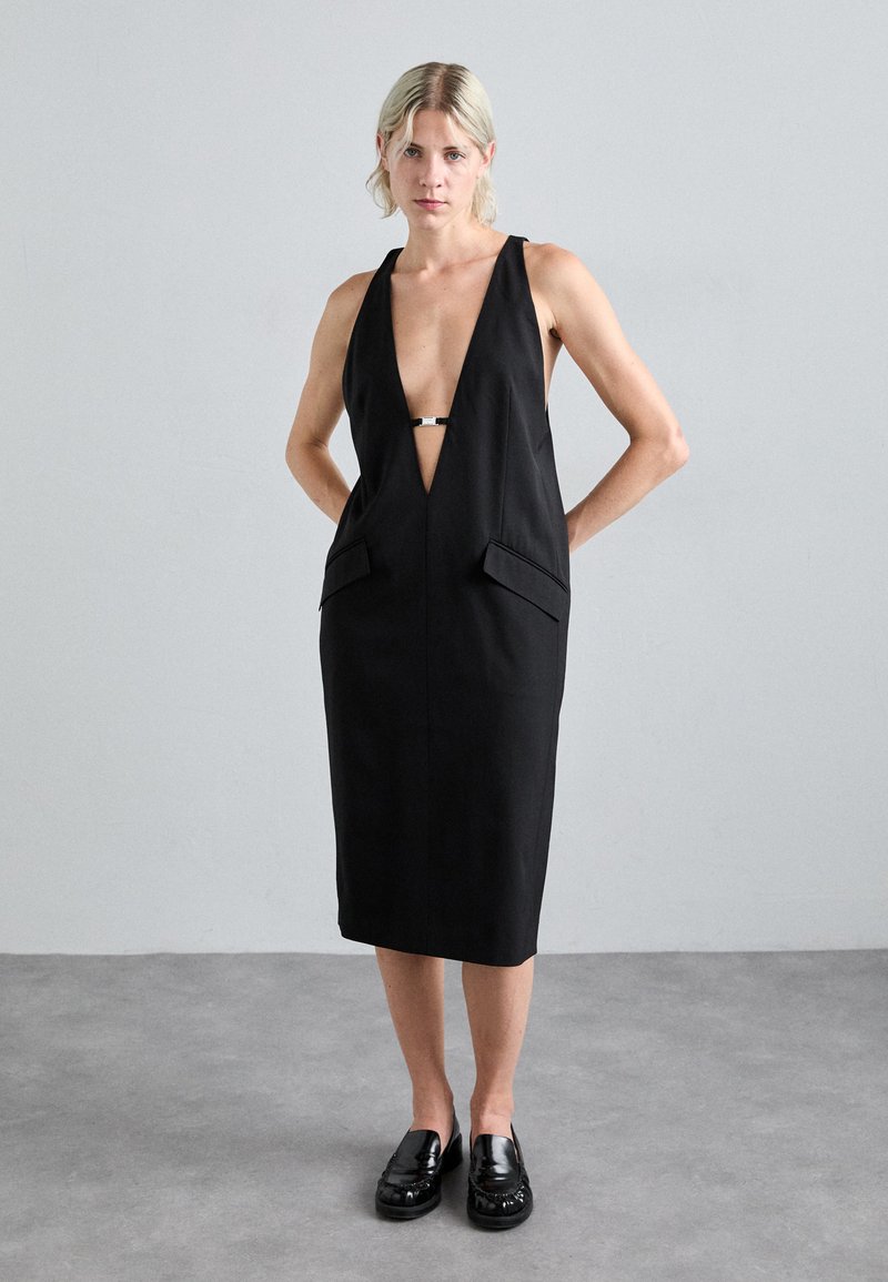Schwarzes Kleid mit tiefem V-Ausschnitt, Schulterträgern, zwei vorderen Taschen und einem silbernen Verschlussdetail an der Brust. Glattes Material, knielang.