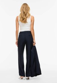 Femme aux longs cheveux blonds portant un haut blanc sans manches, un pantalon large rayé bleu marine, des talons blancs, et tenant une veste assortie.