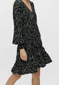 Robe noire à fleurs avec des manches longues, décolleté en V et ourlet volanté. Présente de petits motifs de fleurs blanches et vertes sur un tissu texturé.