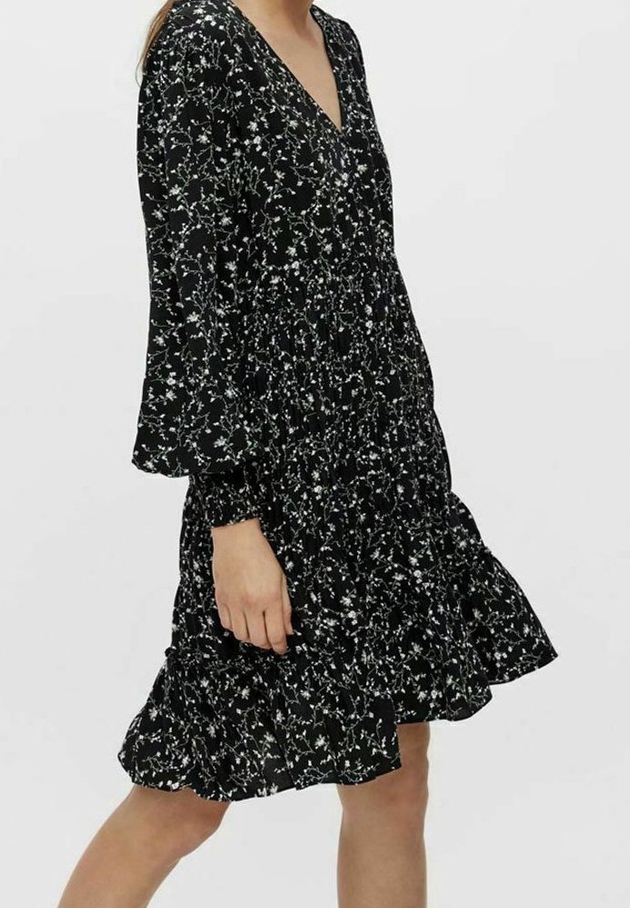 Robe noire à fleurs avec des manches longues, décolleté en V et ourlet volanté. Présente de petits motifs de fleurs blanches et vertes sur un tissu texturé.