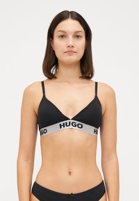 Μαύρο μπραλέτ με τριγωνικό σχήμα, φτιαγμένο από λείο ύφασμα. Διαθέτει μια φαρδιά γκρι ζώνη με την ένδειξη "HUGO" τυπωμένη σε μαύρο στο κάτω μέρος.