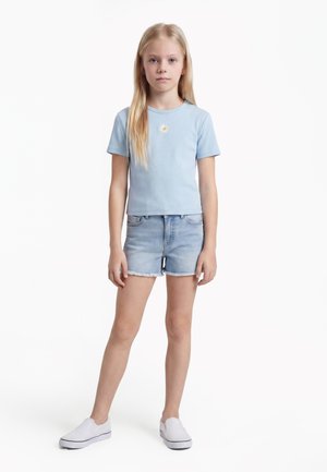 Fiatal lány hosszú szőke hajjal, világoskék margarétás hímzett pólót, farmer rövidnadrágot és fehér bebújós cipőt visel, és előre nézve áll.