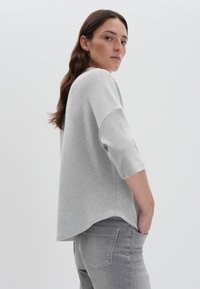 Graues Stricktop mit dreiviertel Ärmeln, strukturiertem Muster und leicht abgerundetem Saum, kombiniert mit grauen Jeans. Minimalistisches Design, nahtlose Akzente.
