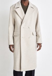 Manteau en laine beige à double boutonnage avec revers, deux poches avant et accents de boutons. Texture lisse et design à la longueur genou.