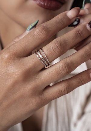 Anillos apilados de plata con texturas trenzadas y lisas, colocados en una mano con arte de uñas en tonos verdes y negros.