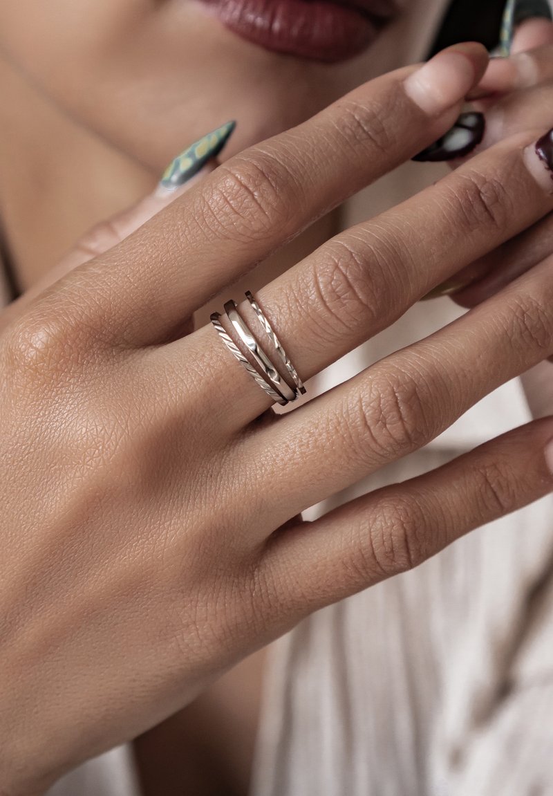 Anillos apilados de plata con texturas trenzadas y lisas, colocados en una mano con arte de uñas en tonos verdes y negros.