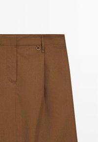Jupe marron taille haute avec passants pour ceinture et un petit anneau métallique près de la taille sur un fond uni.