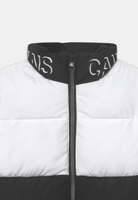 Prošívaná bunda v černo-bílé barevné kombinaci. Má vysoký límec, přední zip a detaily s logem na límci a horní části bundy.