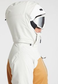 White ski jacket with a brown accent, featuring a hood, zipper, and a streamlined design. Worn with a white ski helmet and goggles.