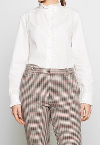 Une chemise blanche à manches longues avec un col ruffle et une patte de boutonnage, associée à un pantalon à carreaux marron avec des accents rouges et noirs.