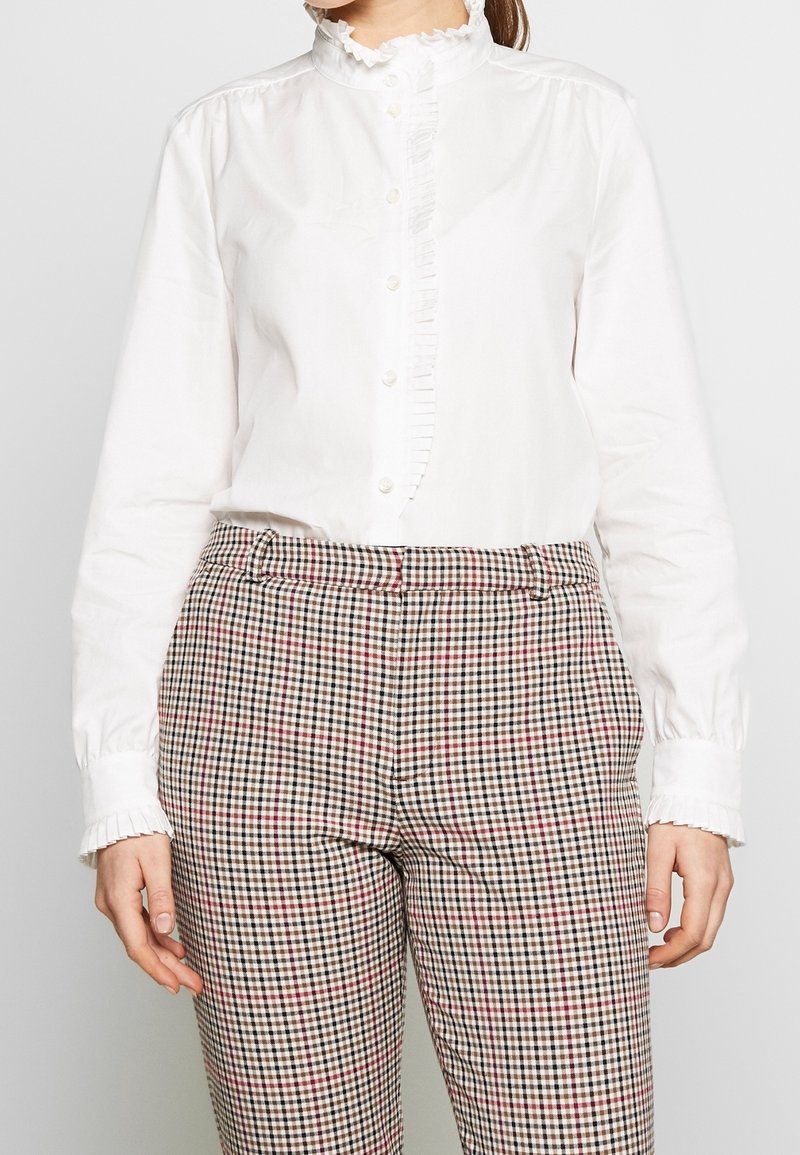 Une chemise blanche à manches longues avec un col ruffle et une patte de boutonnage, associée à un pantalon à carreaux marron avec des accents rouges et noirs.