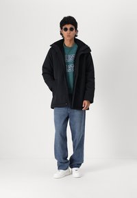 Black parka jacket with a high collar, gray sweatshirt underneath, blue jeans, and white sneakers. Round sunglasses complete the look.