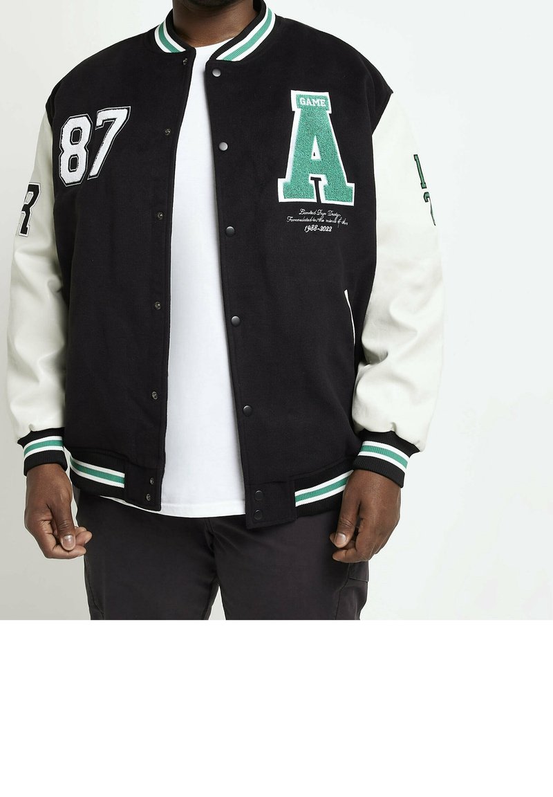 Homme portant une veste varsity noire et blanche avec un col et des poignets rayés vert et blanc, grands écussons "87" et "A" sur le devant.