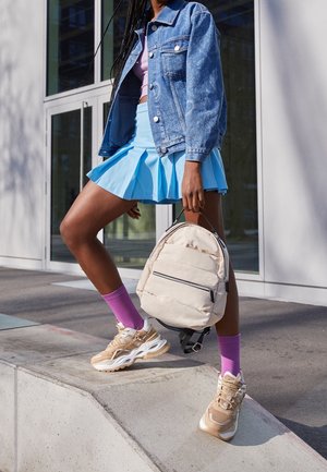 Mujer con chaqueta vaquera, falda plisada azul, calcetines rosas, zapatillas beige, sosteniendo una mochila beige, de pie al aire libre cerca de un edificio con puertas de vidrio.
