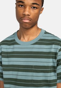 Striped t-shirt in teal and dark green, featuring short sleeves, a crew neck, and a patch pocket with contrasting stitching.