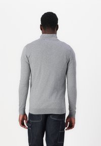 Camisa polo de manga longa cinza com colarinho alto, textura malha, punhos canelados e design ajustado, usada com calças de ganga escuras.